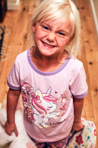 little girl with blond hair and a big smile on her face in purple unicorn glow in the dark pajama set getting ready for bed.