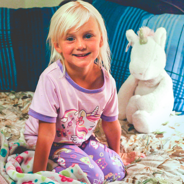 4 year old girl wearing purple glow in the dark unicorn pajama set with stuffed unicorn on bed.